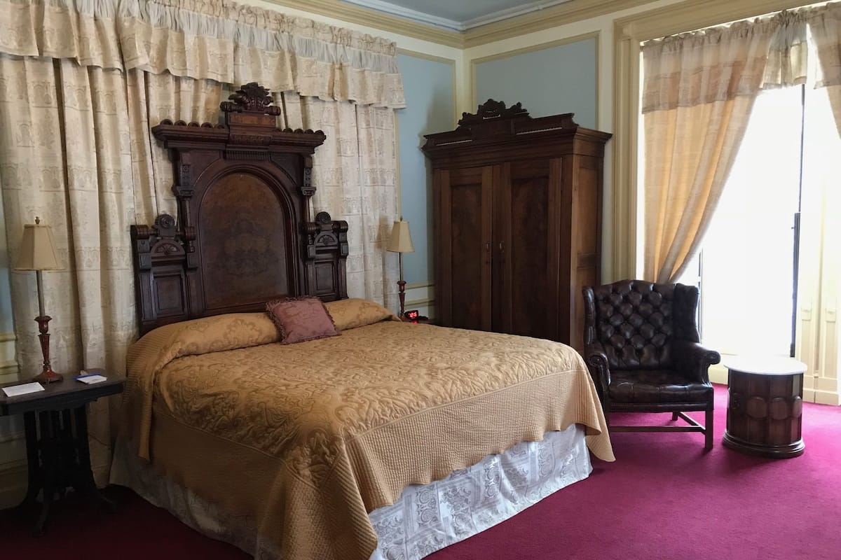 A large bed with a headboard, a wooden wardrobe, a leather armchair, and a small table with a lamp.