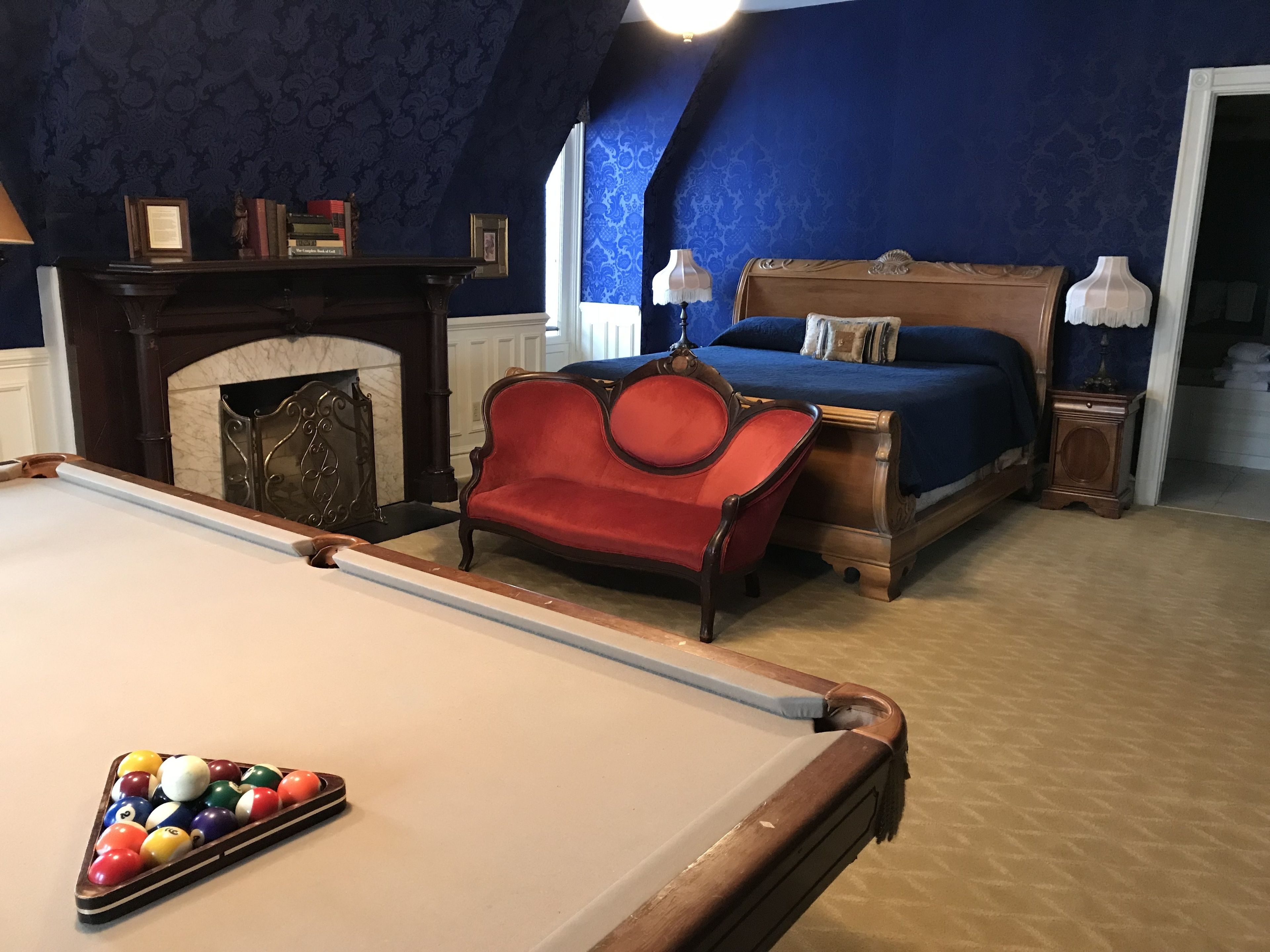 Una habitación con mesa de billar, un sillón rojo, una cama con cubrecama azul y chimenea.