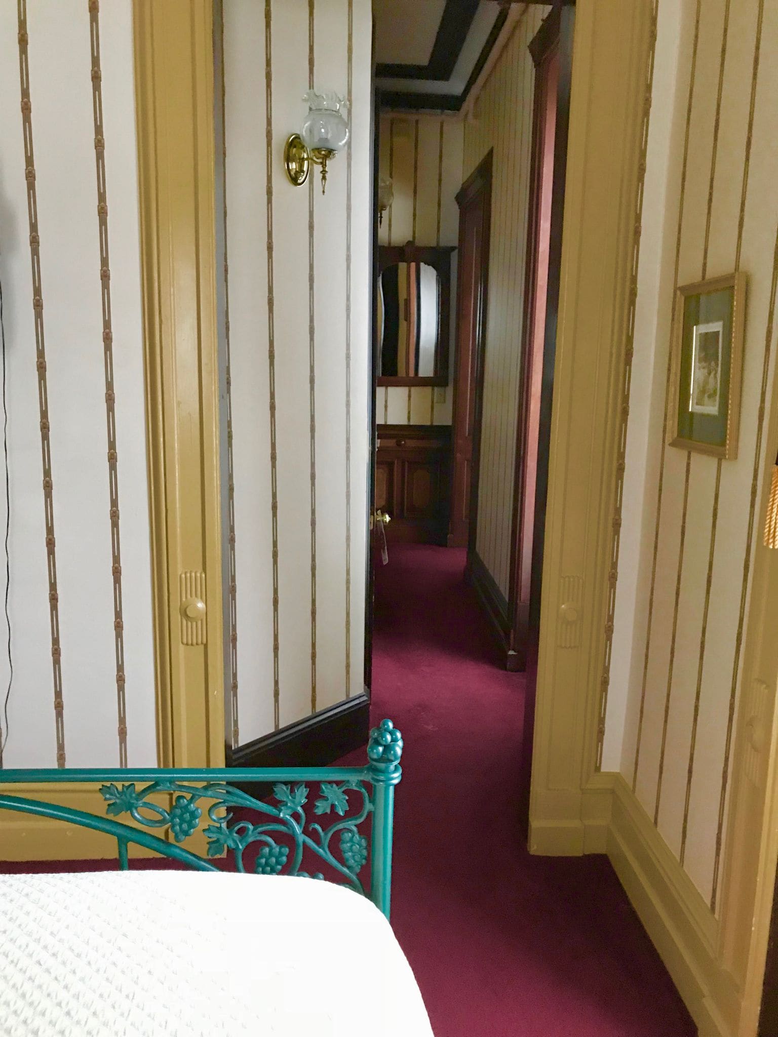 A hotel room with a bed, a mirror, a door, and a hallway with a red carpet.