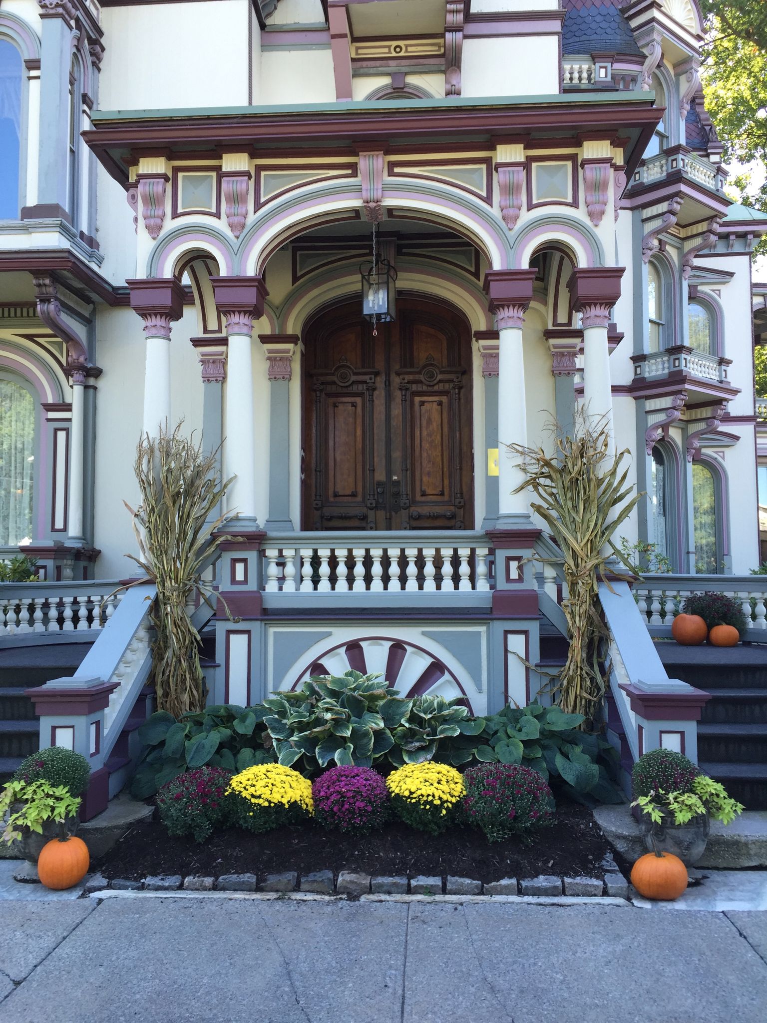 Un edificio de estilo victoriano con una entrada imponente, un arco decorativo y un jardín delantero bien cuidado con diversas plantas y calabazas.