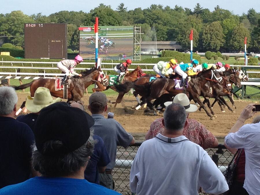 A horse race with jockeys on horses, spectators watching, and a scoreboard in the background.