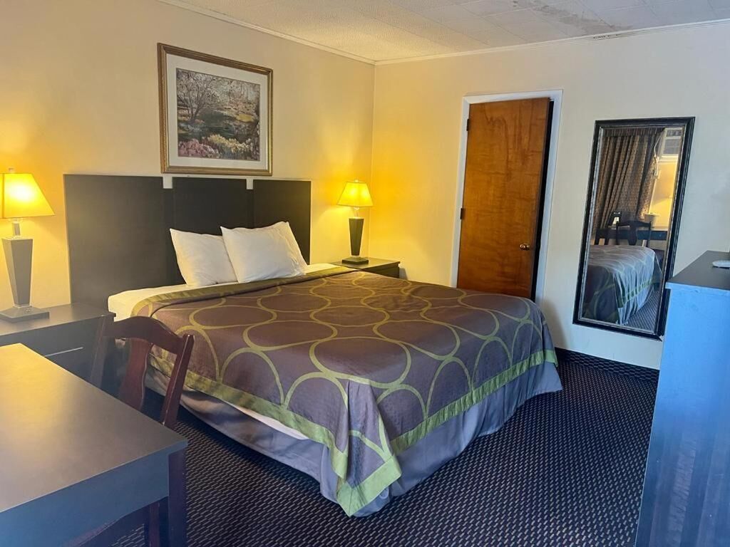 A hotel room with a bed, two bedside lamps, a desk, a chair, a door, and a framed picture on the wall.