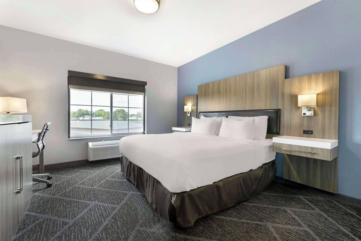 A hotel room with a large bed, a desk, a chair, a window, and a wall-mounted lamp.
