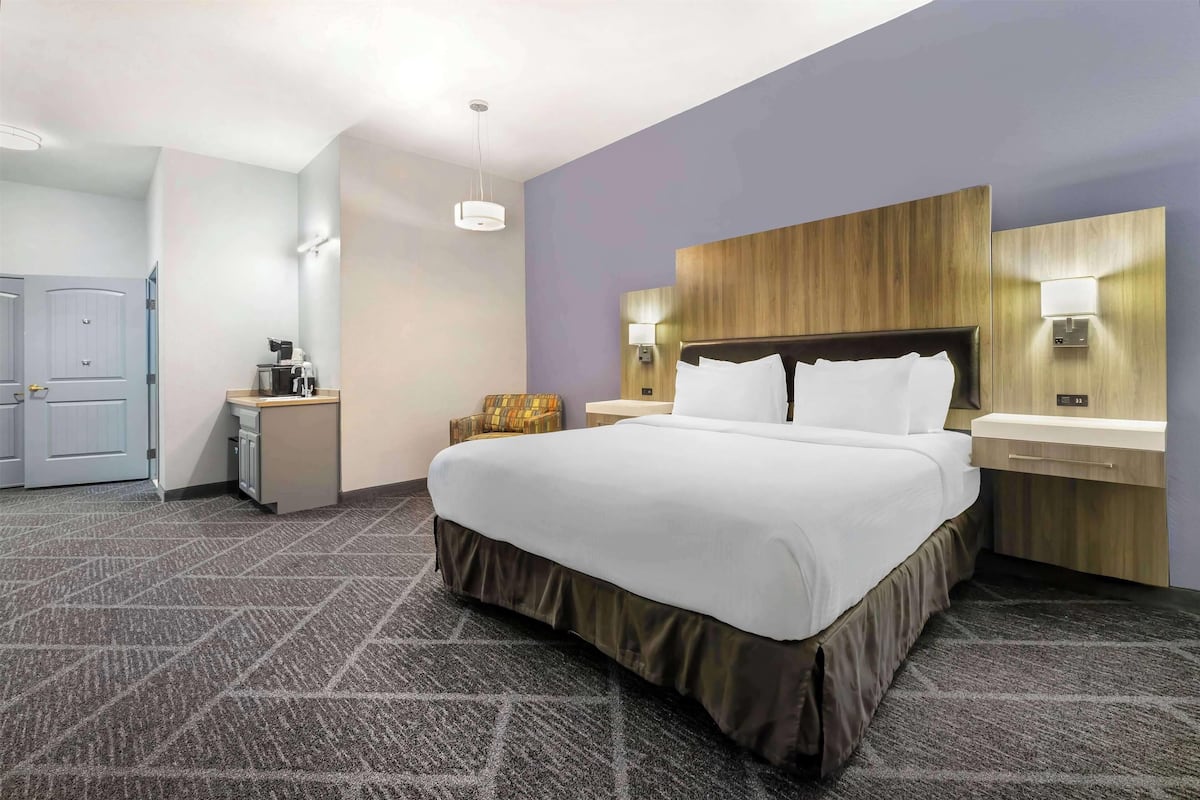 A hotel room with a large bed, a wooden headboard, a bedside table with a lamp, and a small seating area with a chair and a table.