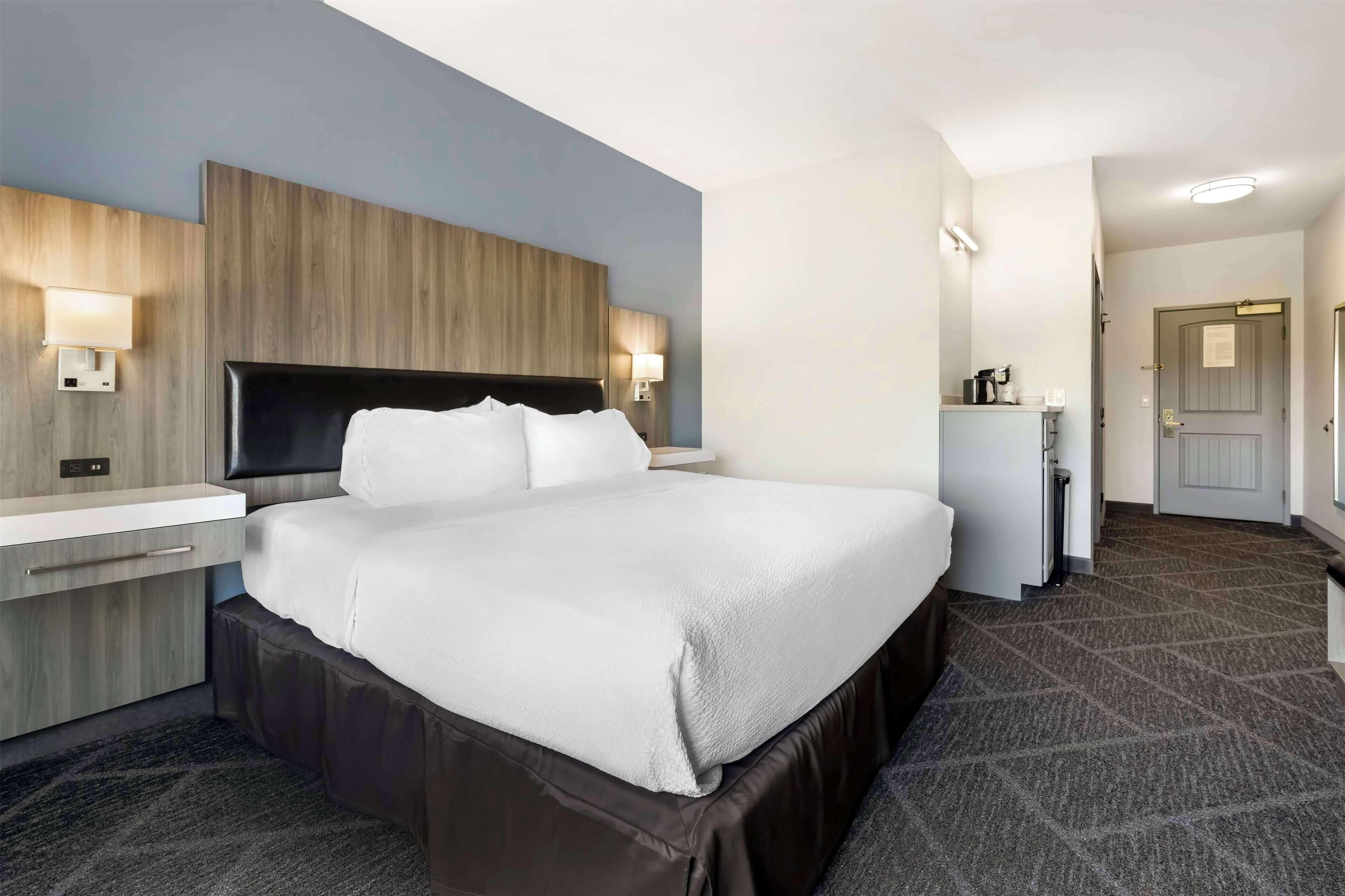 A modern hotel room with a large bed, bedside tables, a built-in desk, and a small kitchenette.
