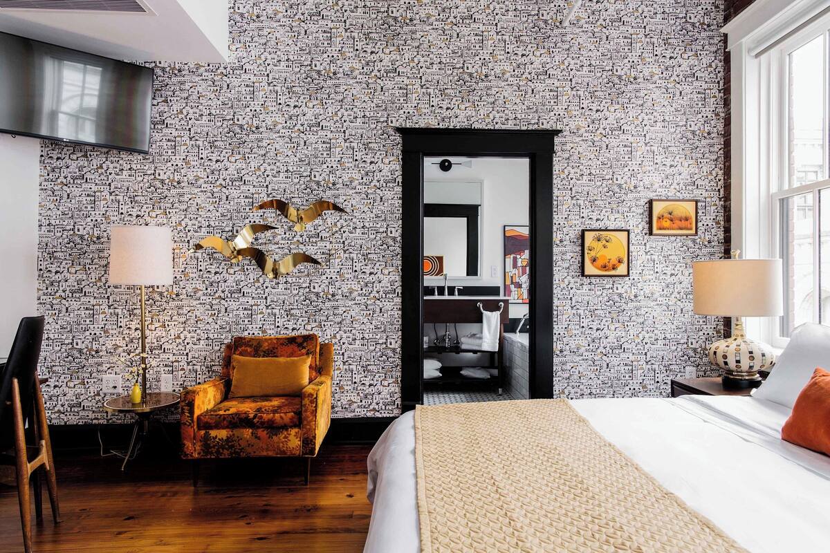 Ein Hotelzimmer mit gemustertem Wandteppich, einem Bett, einem Sofa, einem Sessel, einer Lampe und Bildern an der Wand.