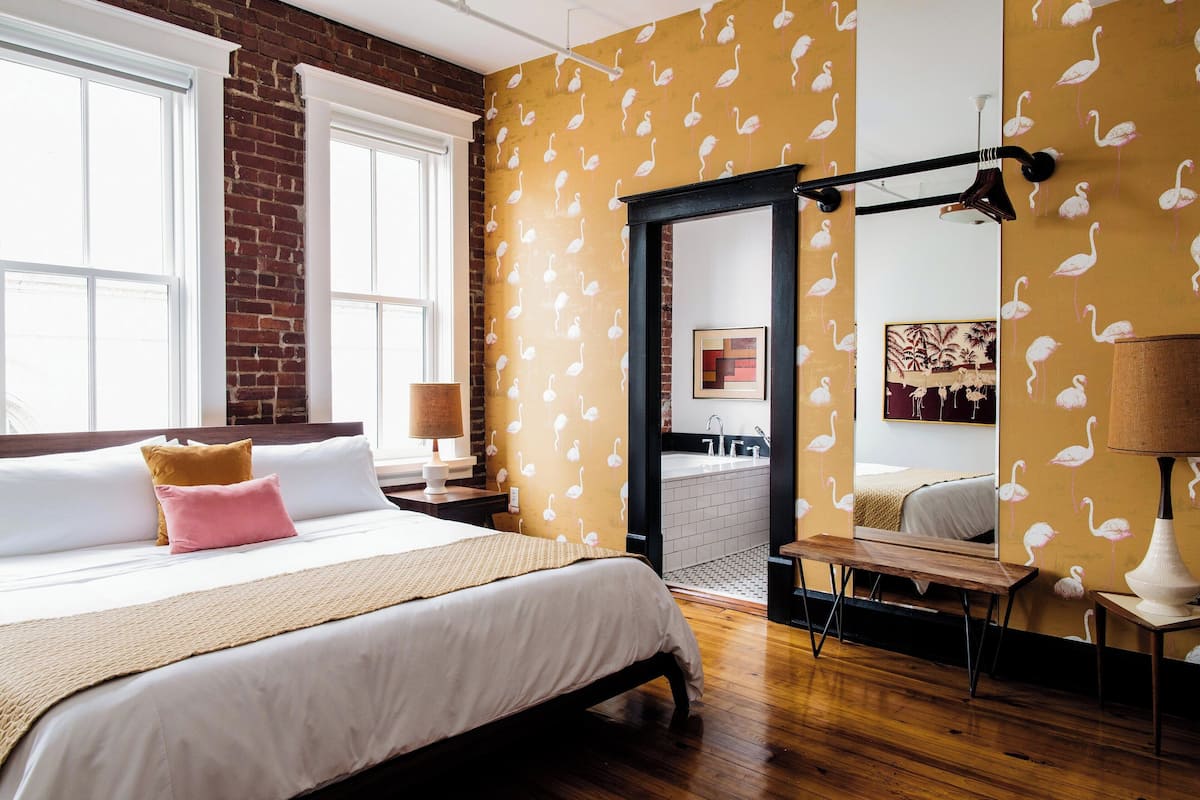 Ein Schlafzimmer mit einem großen Bett, einer hölzernen Bank, einem Spiegel und einem durchgehenden Türrahmen einsehbaren Badezimmer.