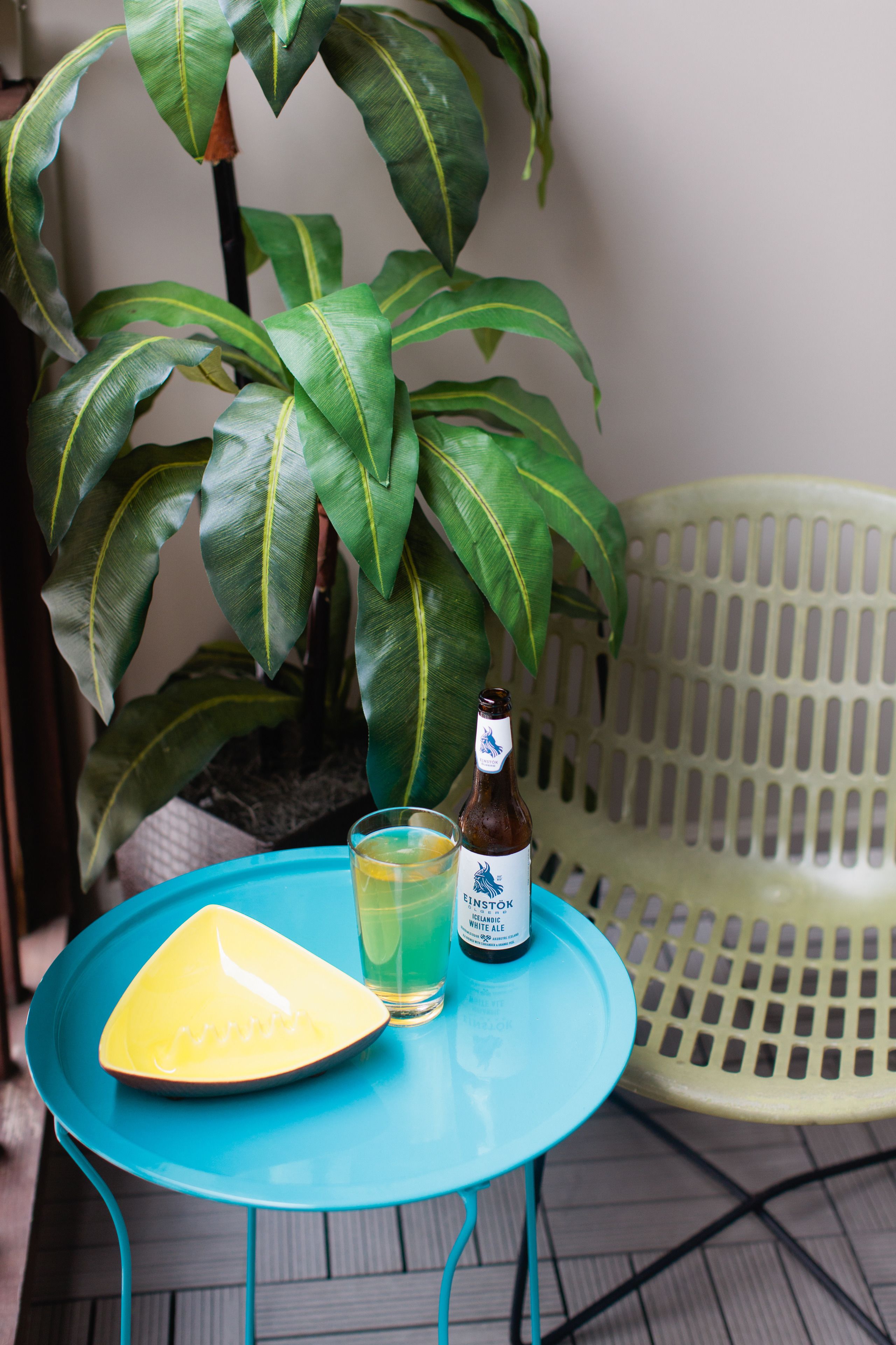 A small table with a bottle of beer and a glass of beer, a yellow plate, and a potted plant.