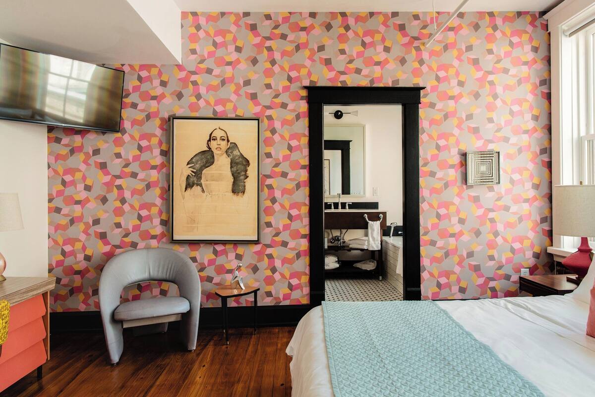 Ein Hotelzimmer mit Wand in geometrischem Muster, einem Bett, einem Sessel, einem kleinen Tisch und einem Spiegel.