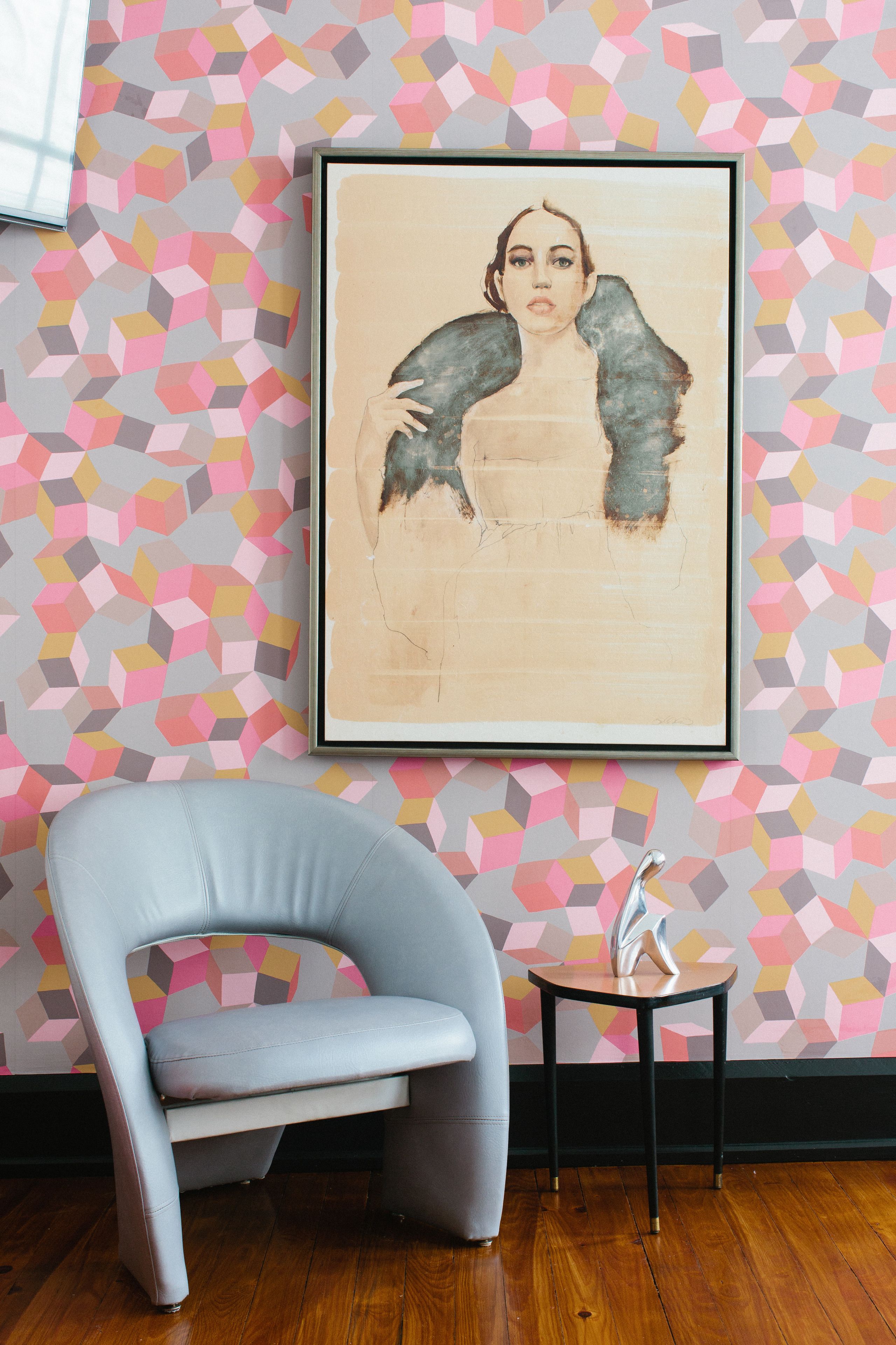 Una obra de arte enmarcada en una pared con un diseño geométrico, una silla moderna y una mesita con un objeto decorativo.