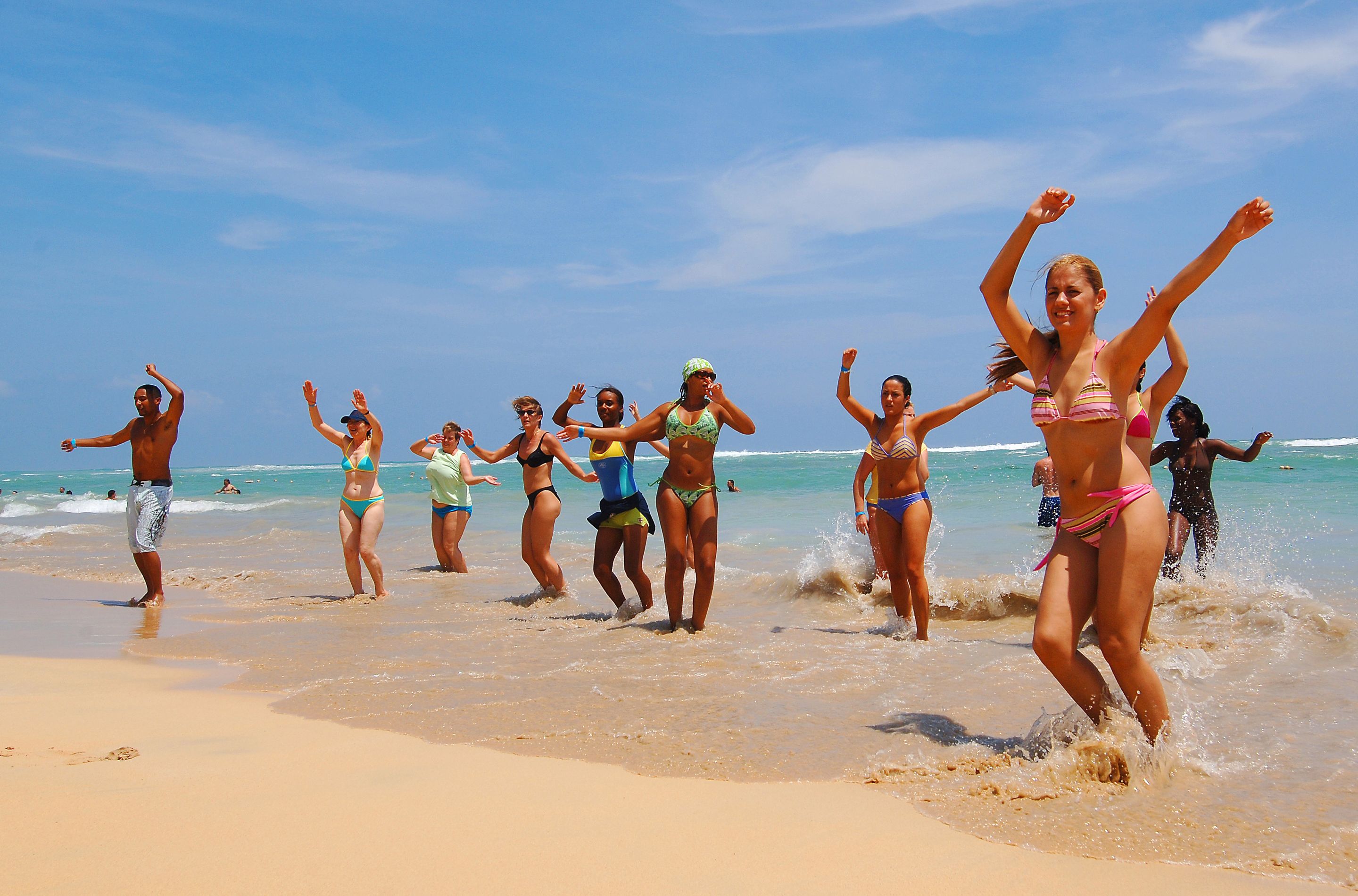 A group of people on a beach, some in swimwear, with waves and a clear sky in the background.