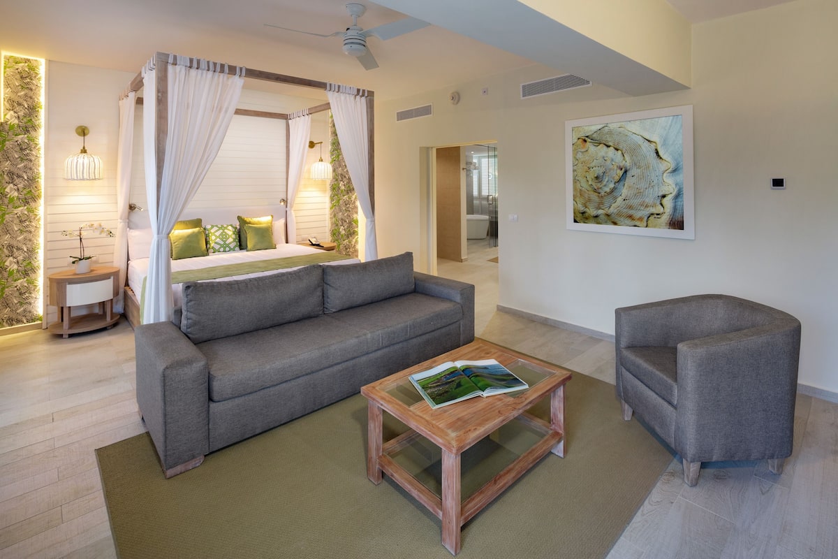 A modern living room with a grey sofa, a wooden coffee table with a magazine, a grey armchair, and a glass door leading to a bathroom.