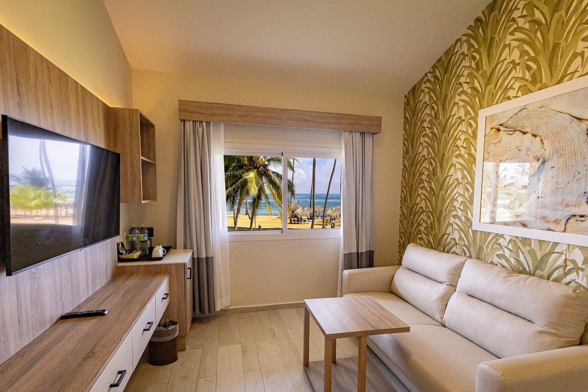 A modern living room with a TV, a sofa, a coffee table, and a view of the beach.