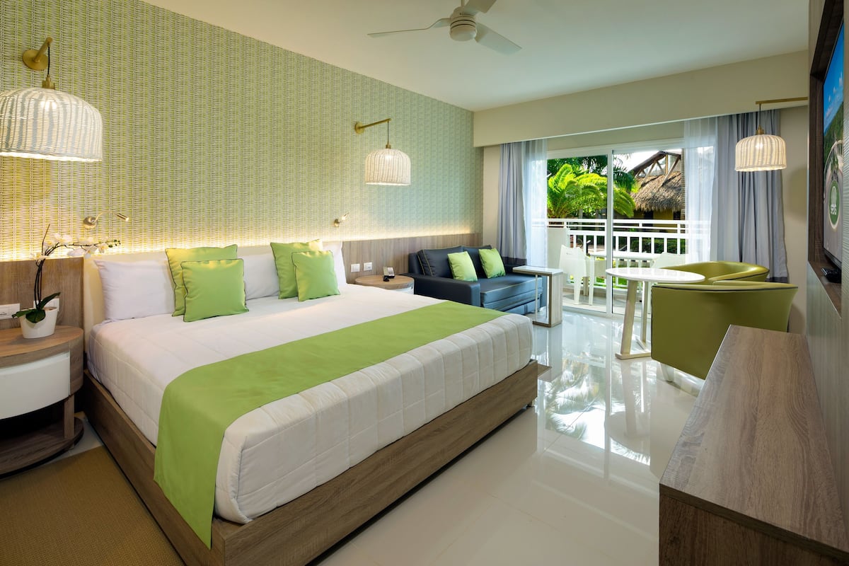 A modern hotel room with a large bed, a balcony with a view of greenery, and a dining area with a table and chairs.