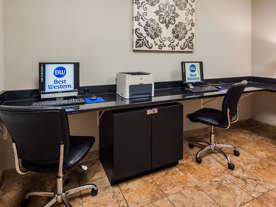 Et hotelarbejdsområde med to computerborde, hvert med en skærm der viser 'Best Western'-branding, en printer og et tastatur. Der er to sorte kontorstole, et skab og en dekorativ vægdekoration.