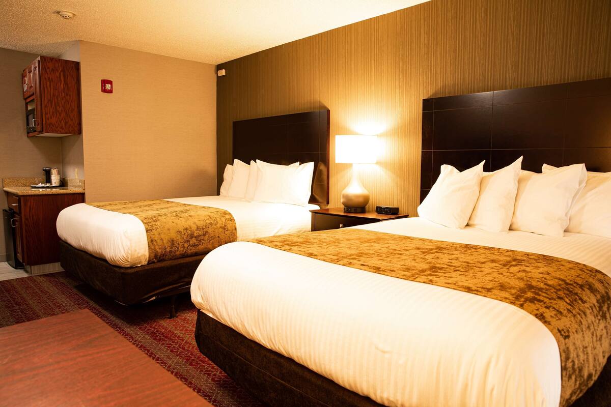 A hotel room with two beds, a wooden headboard, a nightstand with a lamp, and a small desk with a coffee maker.