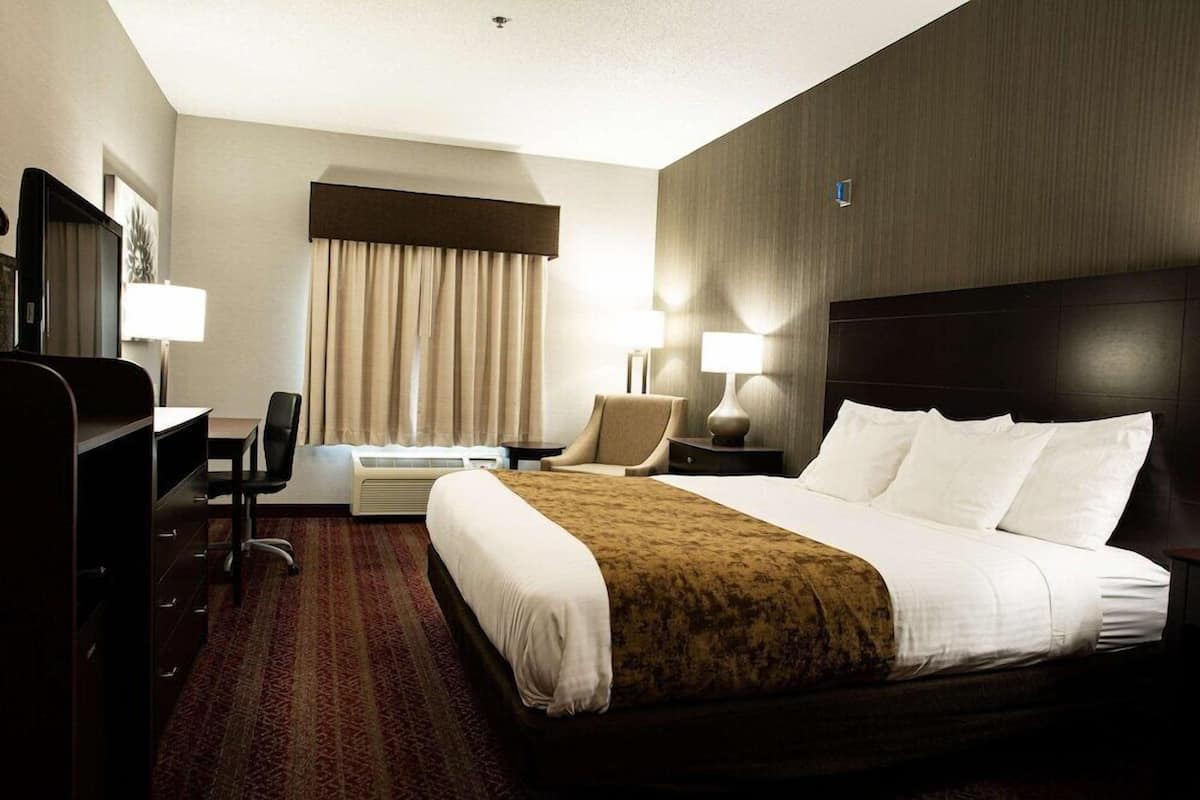 Quarto de hotel com cama, escrivaninha, cadeira e uma janela grande com cortinas.