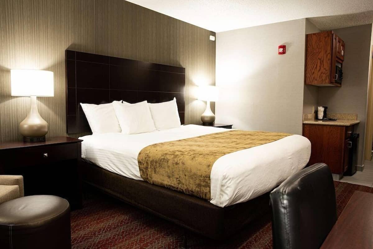 Quarto de hotel com uma cama, duas mesas de cabeceira com luminárias, uma cadeira, uma pequena escrivaninha e uma área de cozinha compacta.
