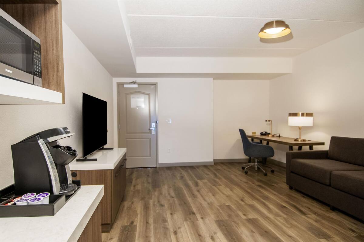 A modern hotel room with a kitchenette, a flat-screen TV, a desk with a chair, a sofa, and a ceiling light.