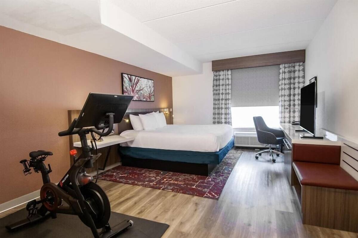 A hotel room with a bed, desk, chair, TV, exercise bike, and a window with curtains.