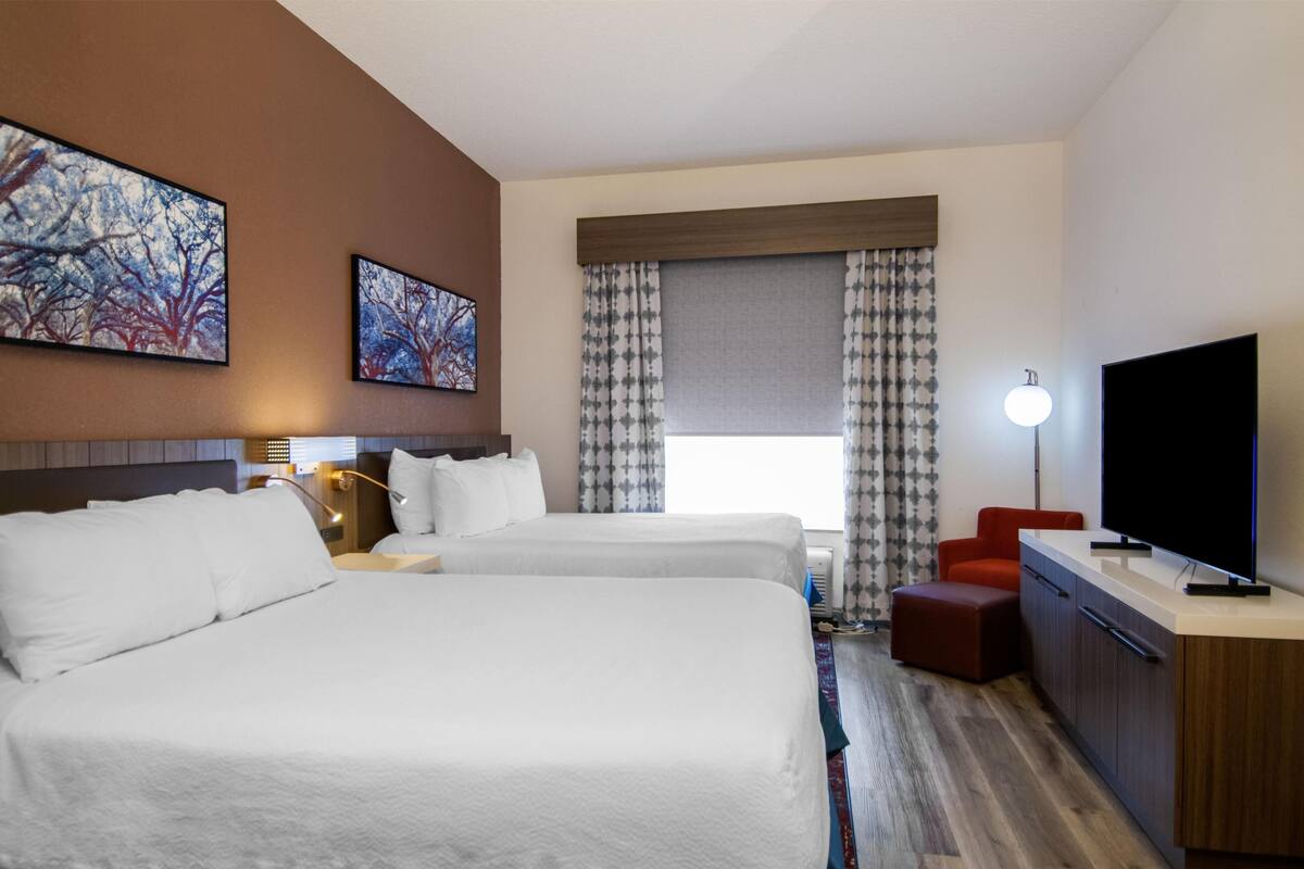 A hotel room with two beds, a TV, a desk, and a window with curtains.