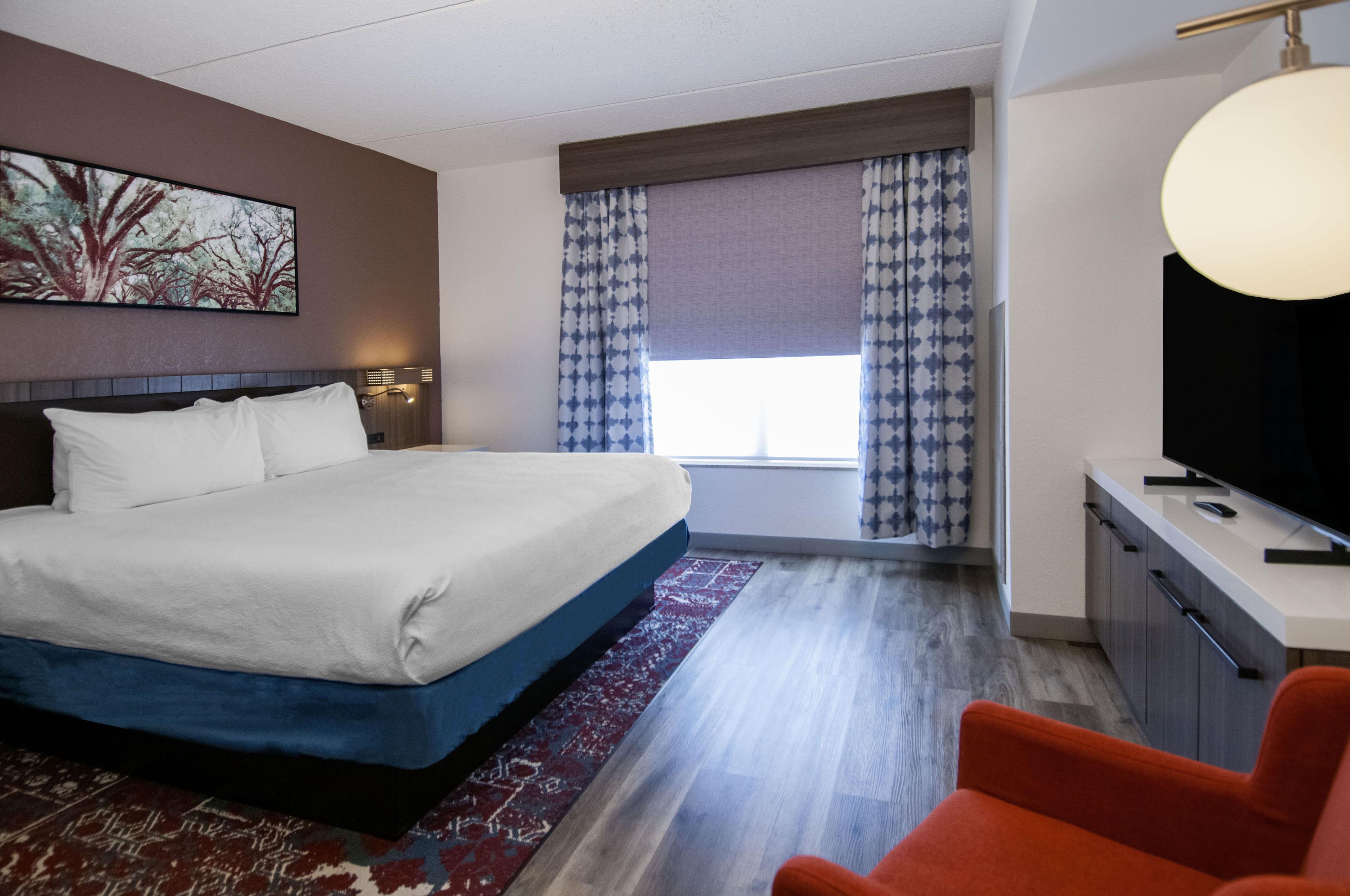 Habitación de hotel con una cama grande, un escritorio con televisor de pantalla plana, una silla roja y una ventana con cortinas estampadas.