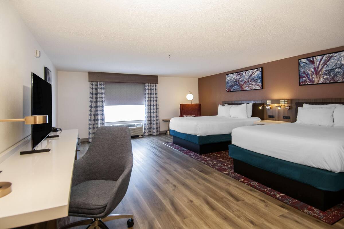 A hotel room with two beds, a desk with a chair, a flat-screen TV, and artwork on the walls.