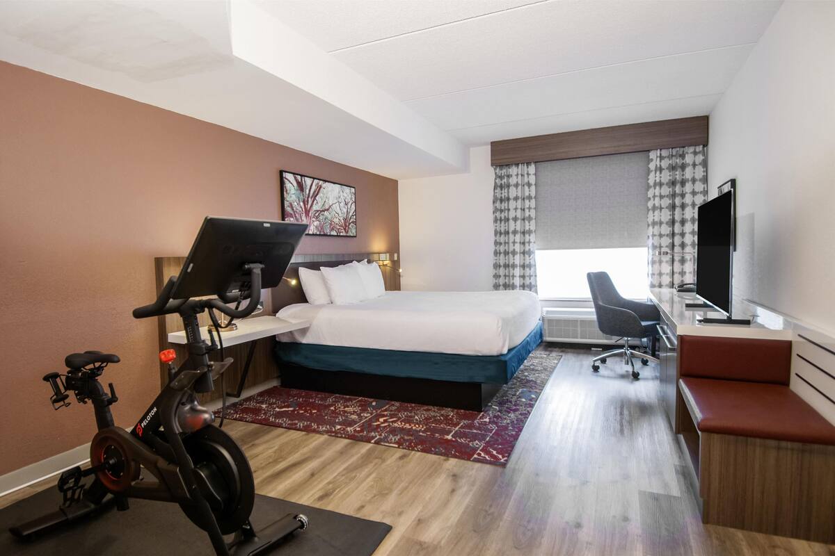 A hotel room with a bed, a desk with a computer, a TV, an exercise bike, and a window with curtains.