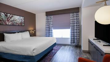 Habitación de hotel con una cama grande, un escritorio con televisor, una ventana con cortinas estampadas y un cuadro en la pared.