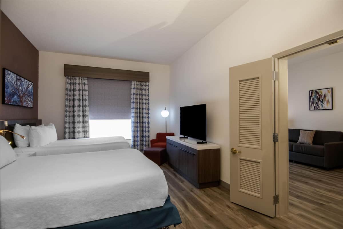 A hotel room with a bed, a desk with a TV, a red chair, a sofa, and a door.