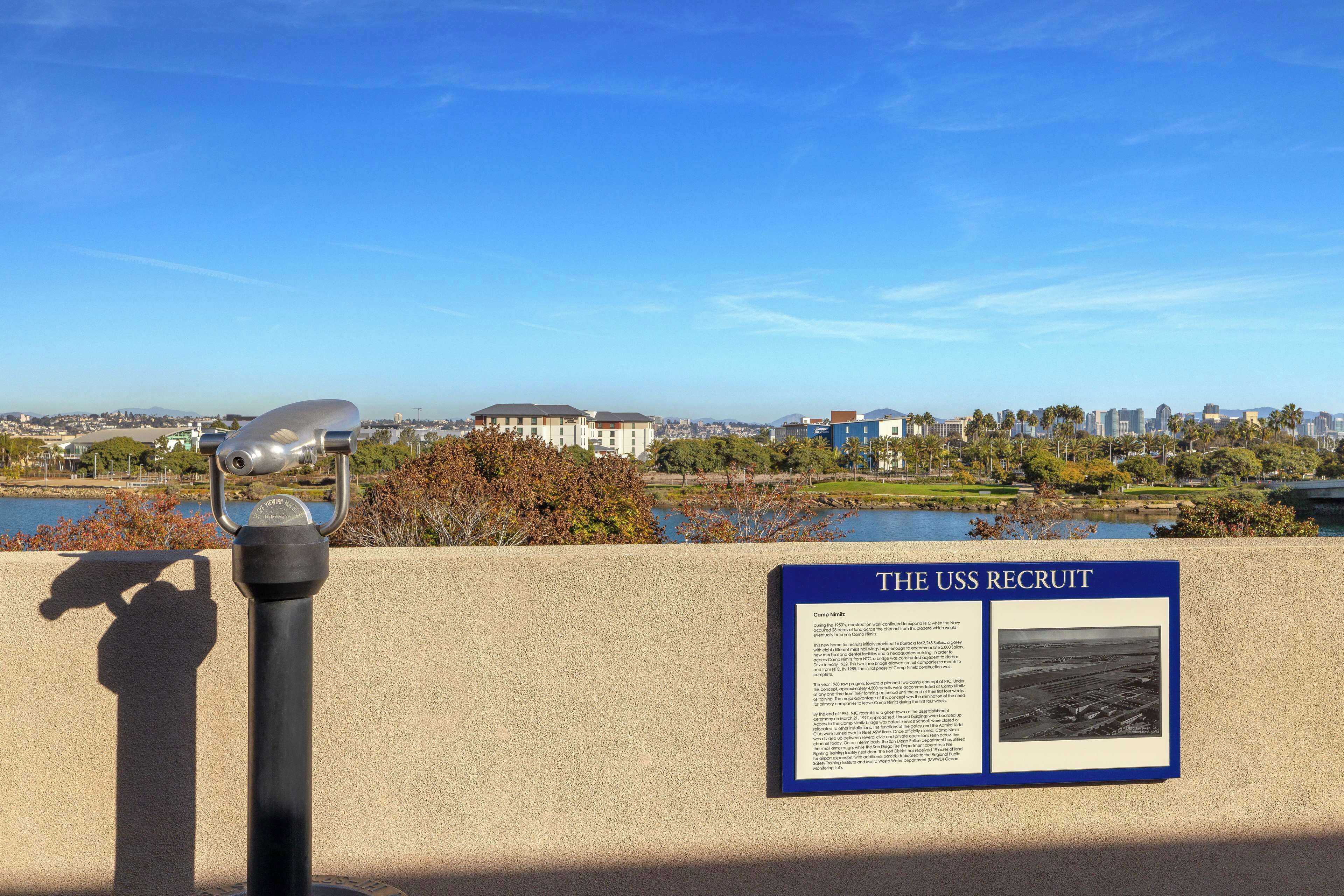 A historical marker titled "THE USS RECRUIT" with a view of a cityscape and a body of water in the background.