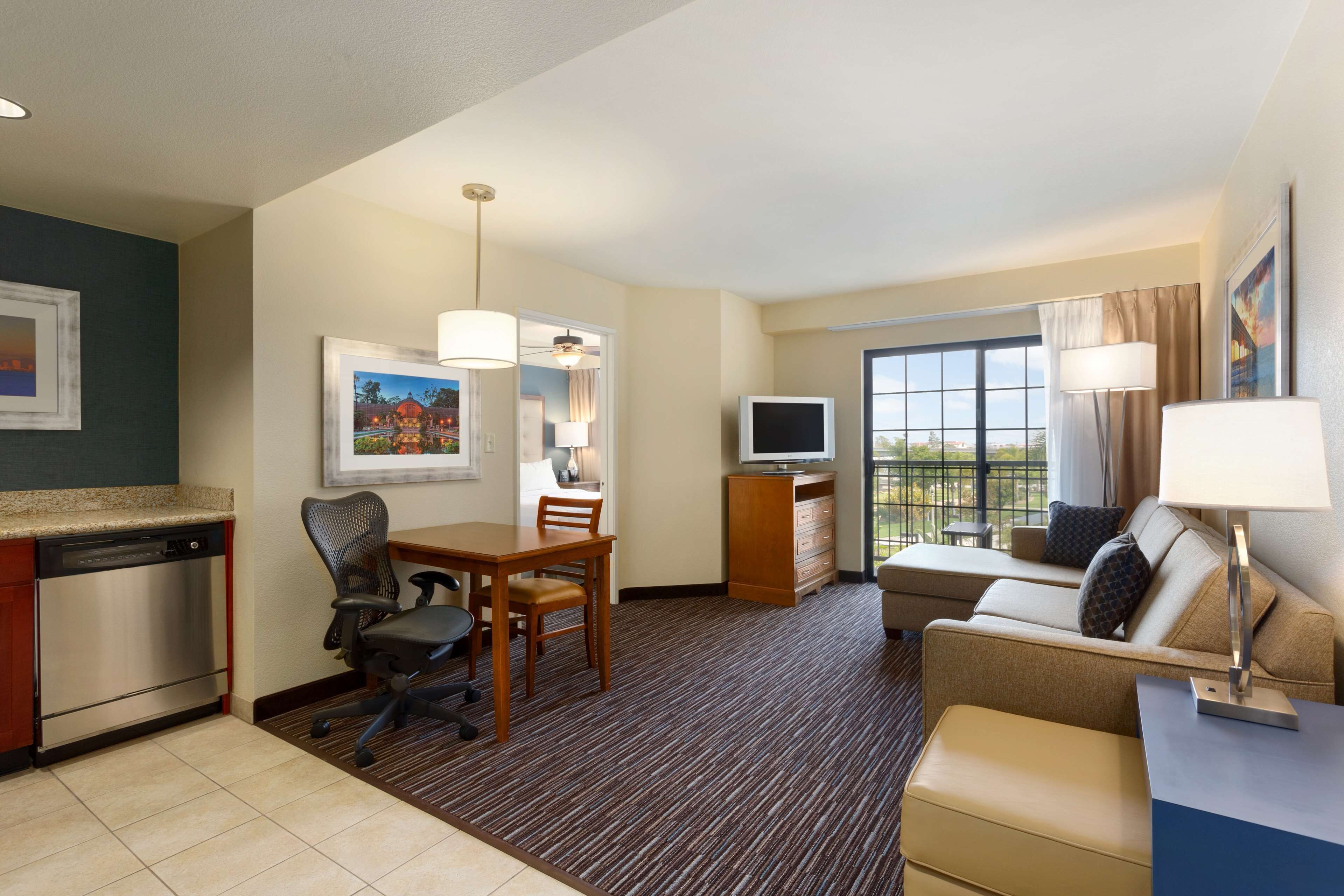 A hotel room with a kitchenette, a dining area, a sofa, a TV, and a large window with a view.