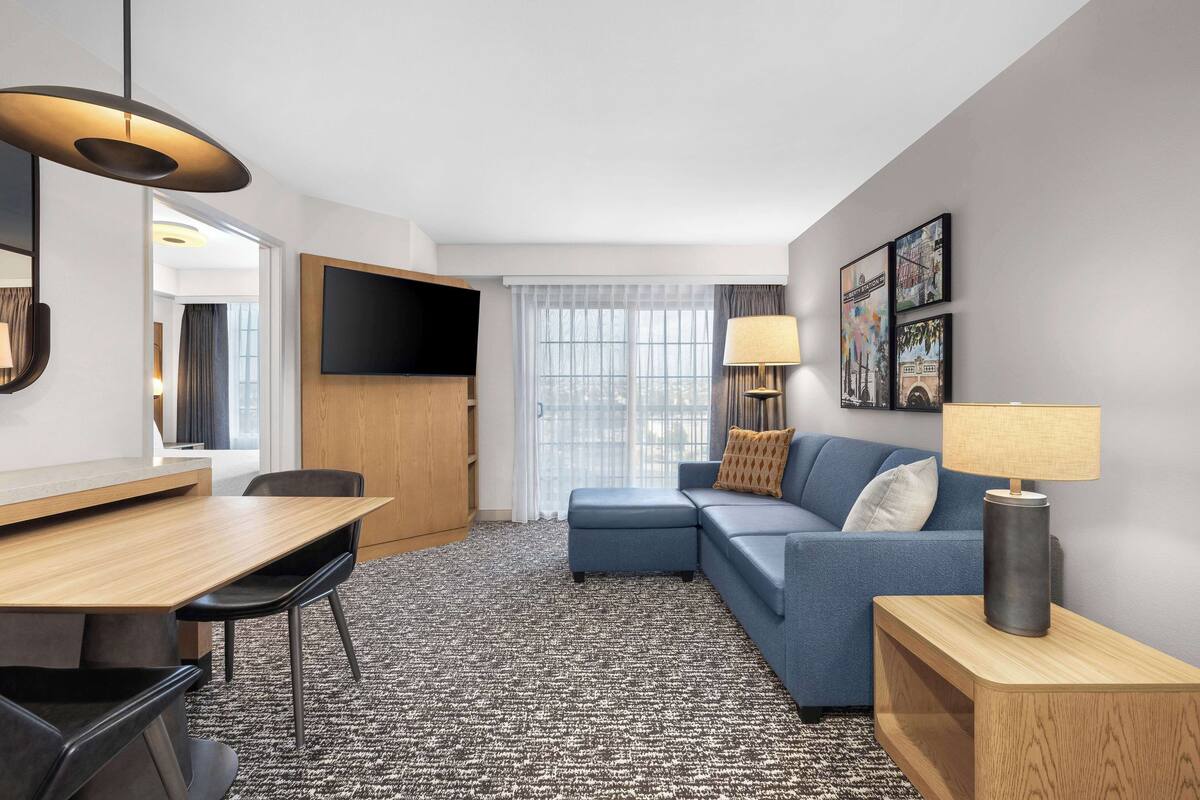 Une chambre d’hôtel moderne avec une table à manger en bois, un canapé bleu, un téléviseur fixé au mur et une moquette à motifs.