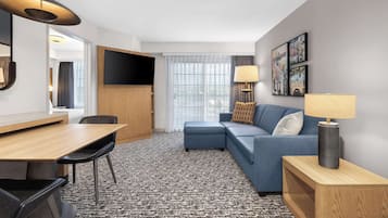 A modern hotel room with a wooden dining table, a blue sofa, a TV mounted on the wall, and a patterned carpet.