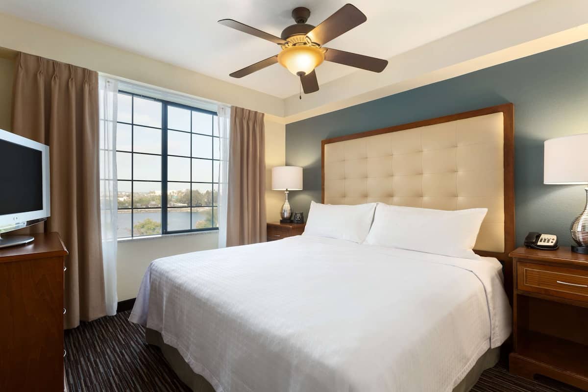 Une chambre d’hôtel équipée d’un lit, d’un ventilateur de plafond, d’une télévision, d’une fenêtre avec des rideaux et d’une table de chevet avec une lampe et un téléphone.