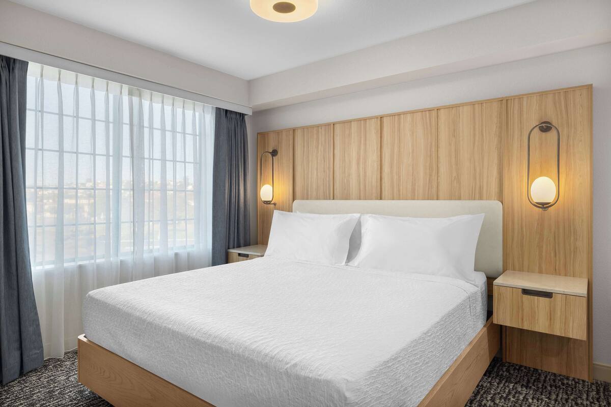 Une chambre d’hôtel avec un grand lit, une tête de lit en bois, deux tables de chevet et une vue sur la ville par la fenêtre.