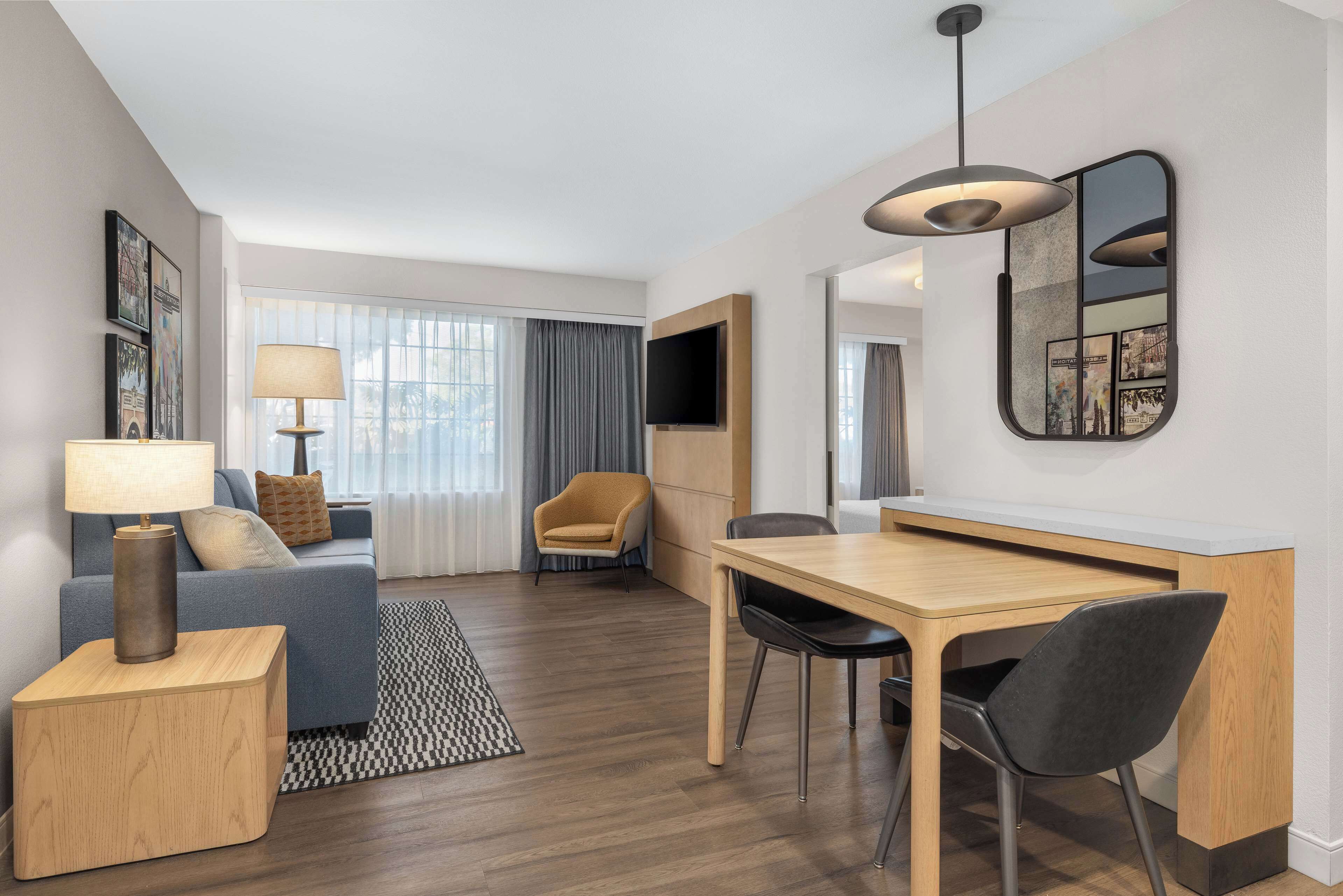 Moderni hotellihuone, jossa on sohva, ruokapöytä tuoleineen, seinälle asennettu televisio ja suuri ikkuna, jossa on verhot.