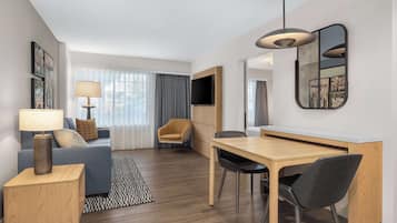 Moderni hotellihuone, jossa on sohva, ruokapöytä tuoleineen, seinälle asennettu televisio ja suuri ikkuna, jossa on verhot.