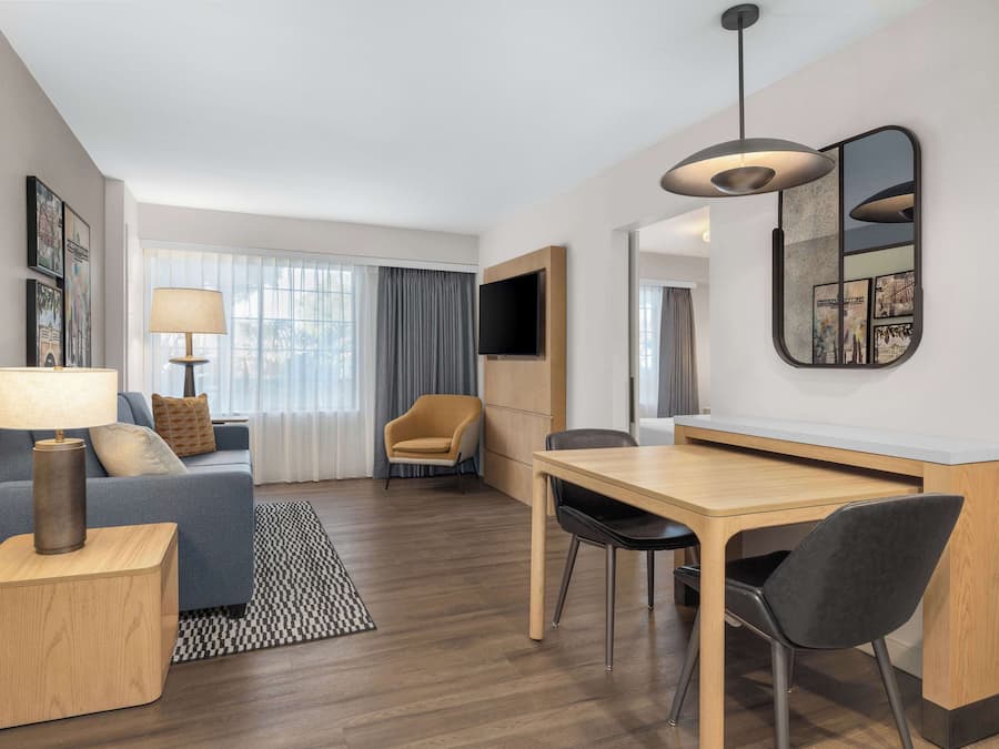 Moderni hotellihuone, jossa on sohva, ruokapöytä tuoleineen, seinälle asennettu televisio ja suuri ikkuna, jossa on verhot.