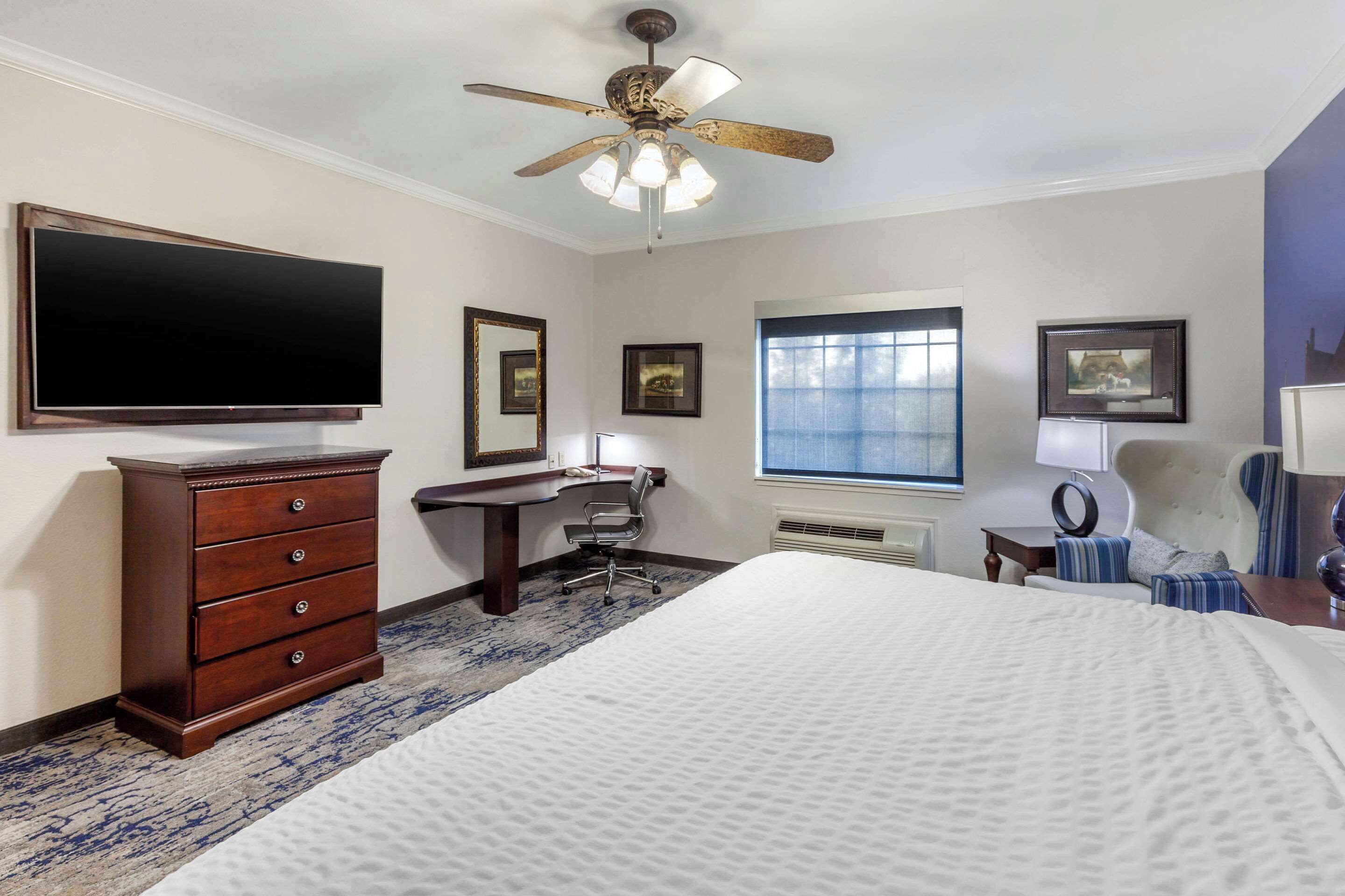 A bedroom with a large bed, a desk, a chair, a dresser, a ceiling fan, and a TV.