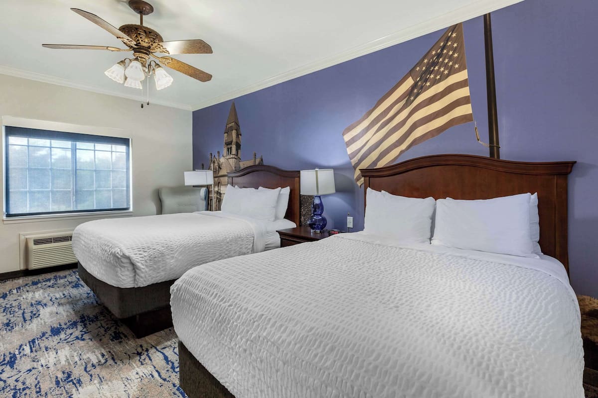 A hotel room with two beds, a ceiling fan, a large window, and an American flag.