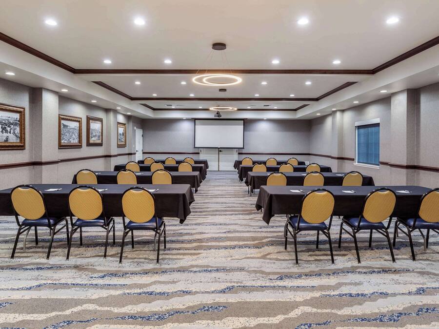 Un salón de conferencias con filas de mesas y sillas, una pantalla de proyector y cuadros en las paredes.