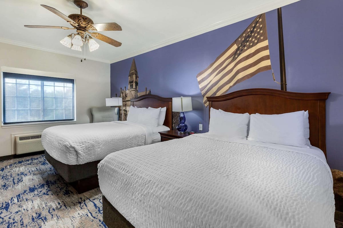 A hotel room with two beds, a ceiling fan, a large window, and an American flag.