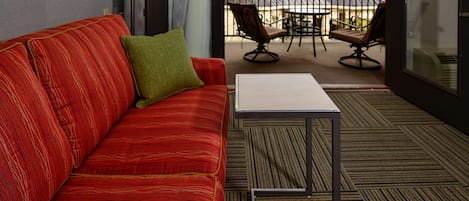 A red sofa with a green pillow, a small white table, and a view of a balcony with chairs and a table.