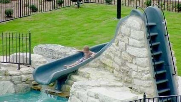 A water slide at a poolside area with a stone structure, surrounded by a metal fence. - Hampton Inn & Suites Fredericksburg (Fredericksburg)