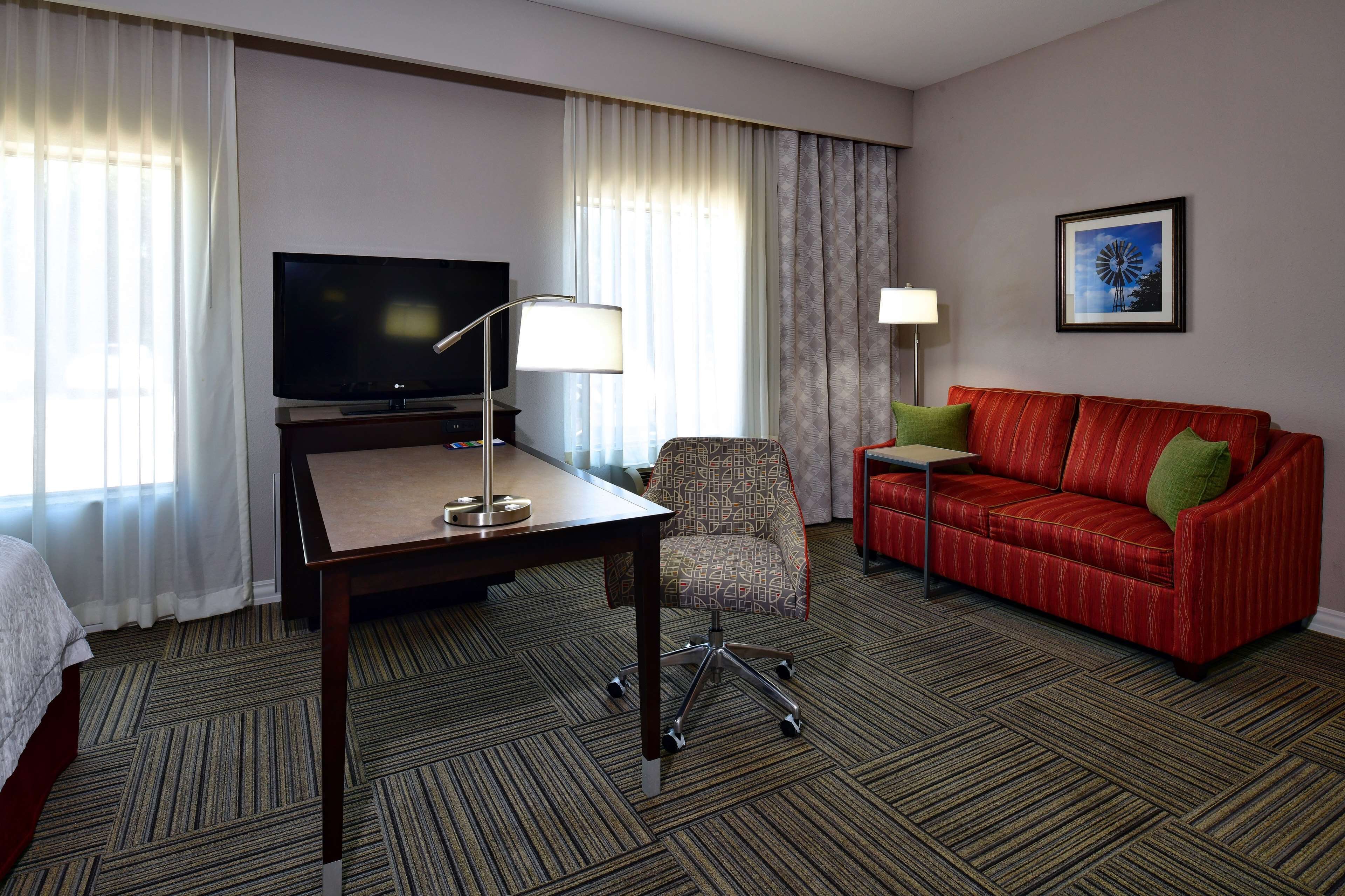 A hotel room with a large flat-screen TV, a desk with a lamp, a red sofa with green cushions, and a patterned armchair.