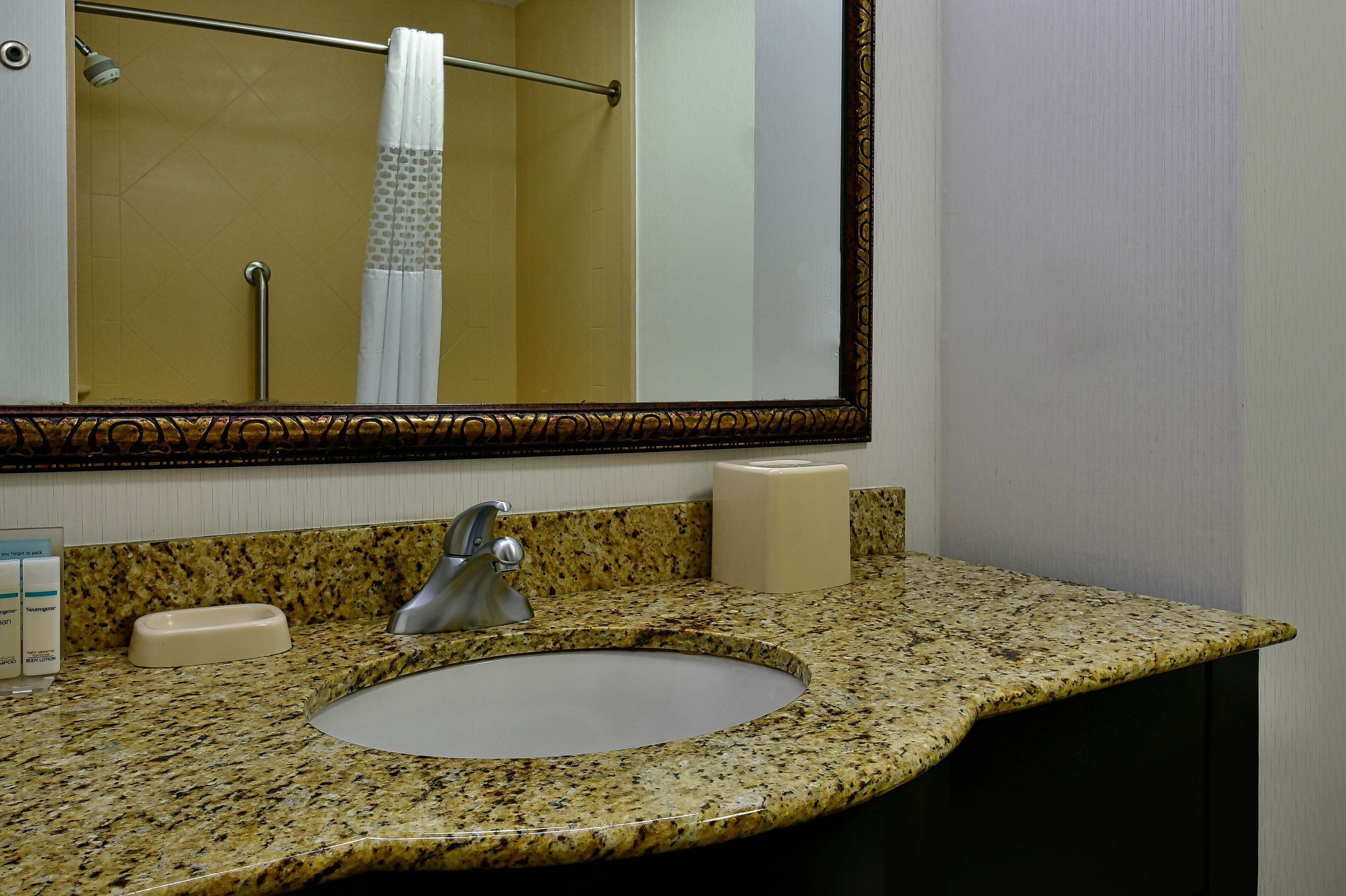 A bathroom with a granite countertop, a sink, a mirror, a towel rack, and a toilet paper dispenser.