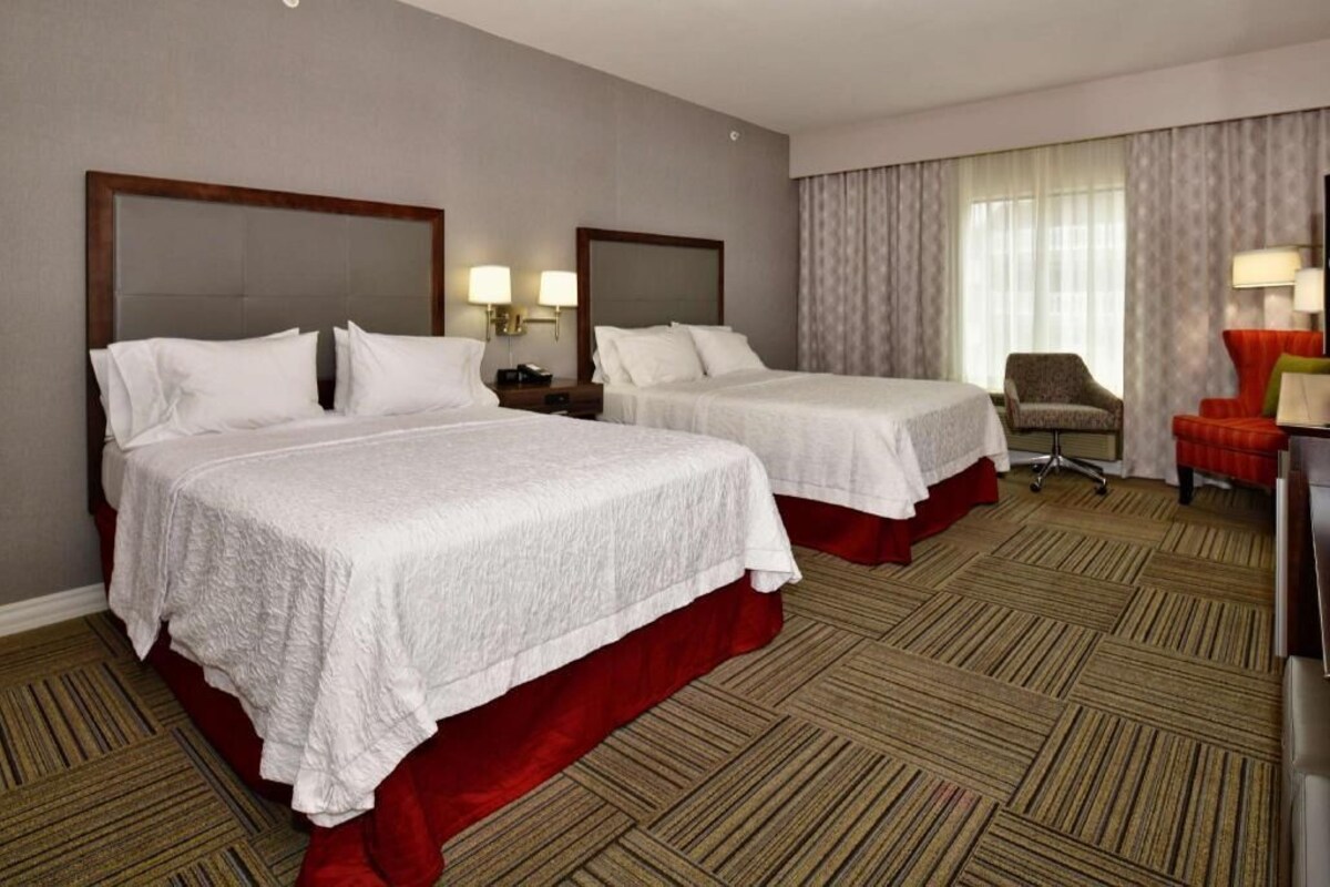 A hotel room with two beds, a desk, a chair, and a large window with curtains.