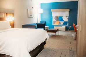 A hotel room with a bed, a desk, a chair, a lamp, and a window with a geometric pattern.