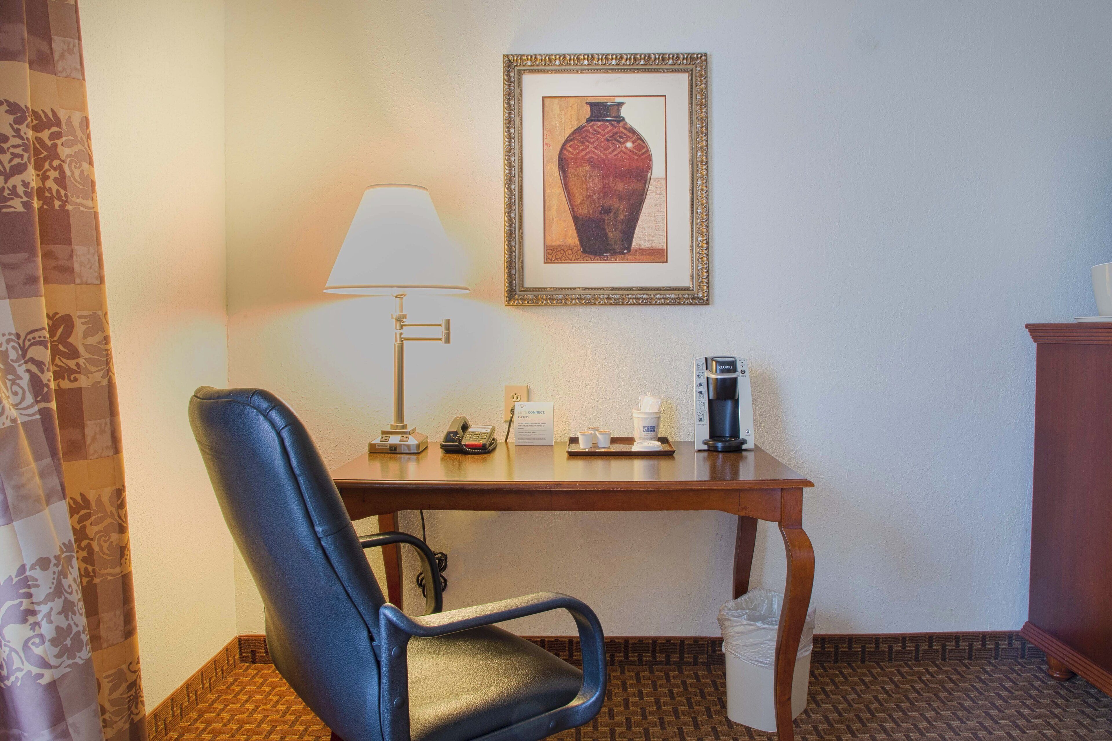 A hotel room with a desk, a chair, a lamp, a framed picture, a coffee maker, and a trash bin.